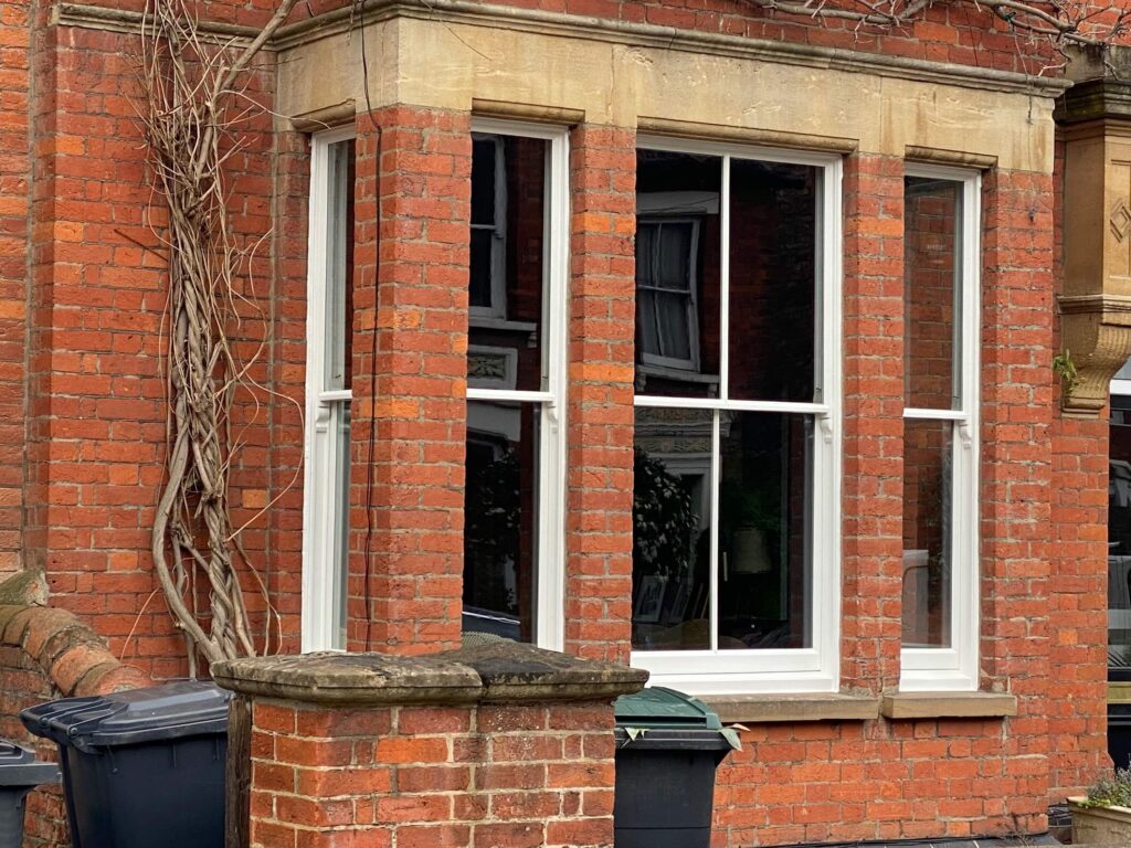 A red brick house with a bay window, white sash windows Cambridge style, a stone window ledge, and a few rubbish bins on the pavement.