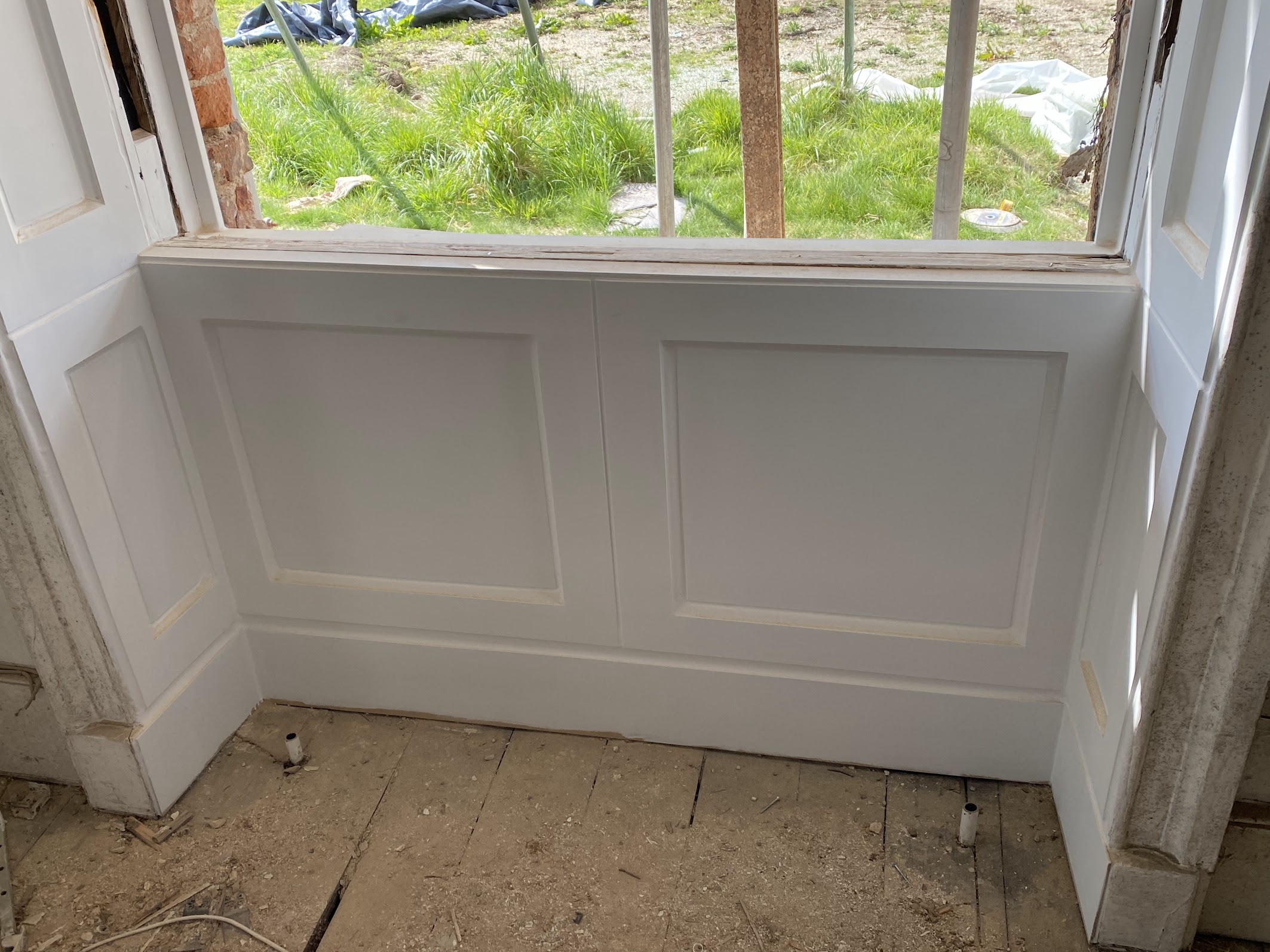 White wooden panelling installed below a sash window in a partially renovated room, with unfinished flooring and construction materials visible outside—perfect for those seeking sash windows Cambridge style.