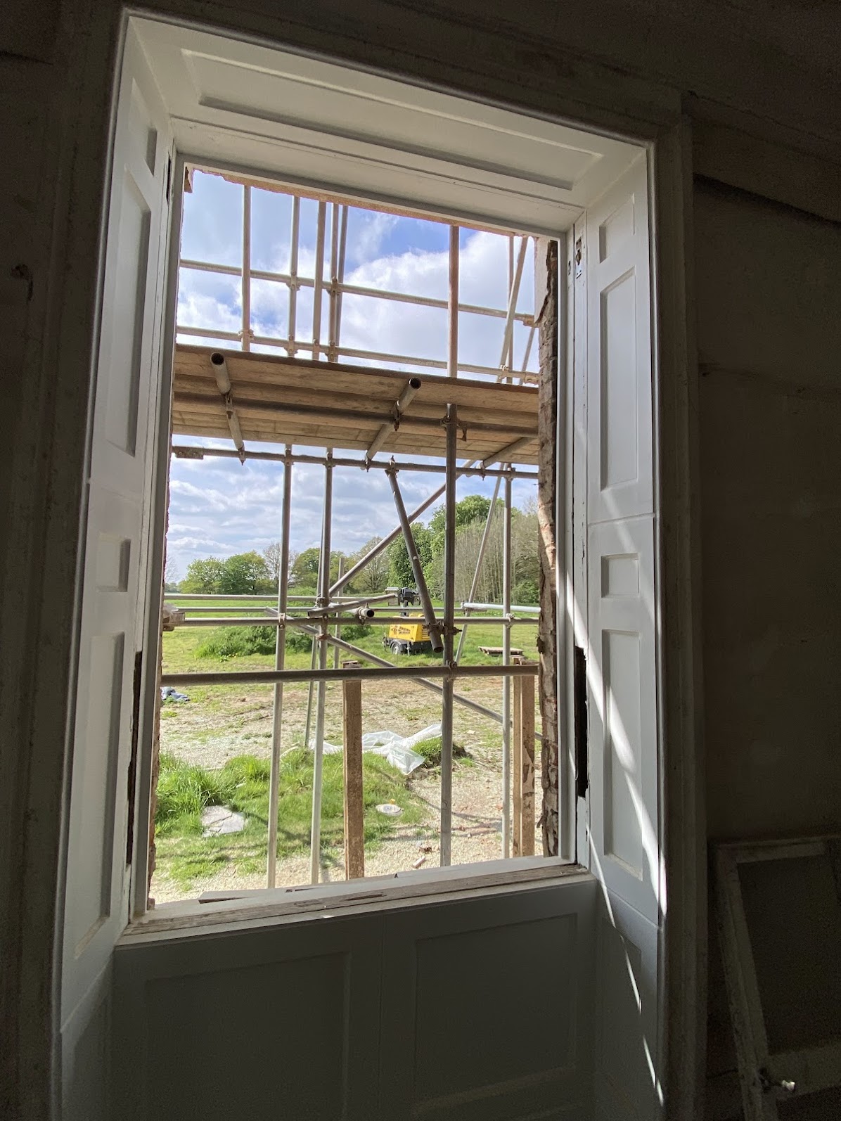 Open sash windows Cambridge with white shutters reveal construction scaffolding outside, overlooking a grassy area and trees beneath a partly cloudy sky.