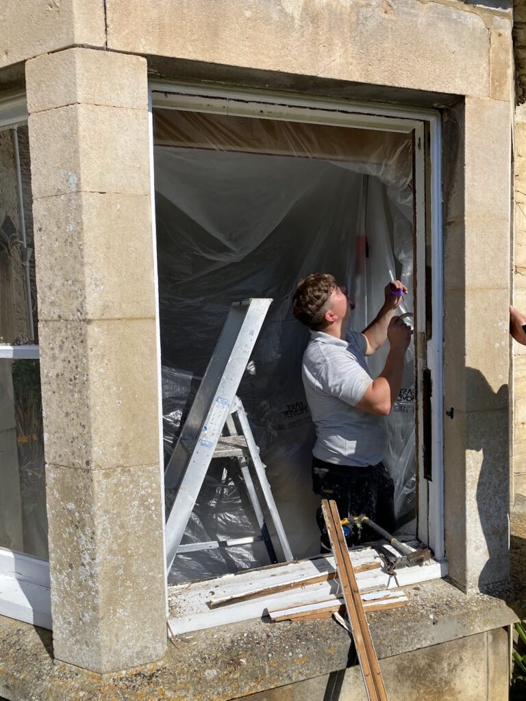 A person uses a tool to work on the window frame of a stone building with sash windows in Cambridge; a ladder and plastic sheeting are set up inside the window opening.