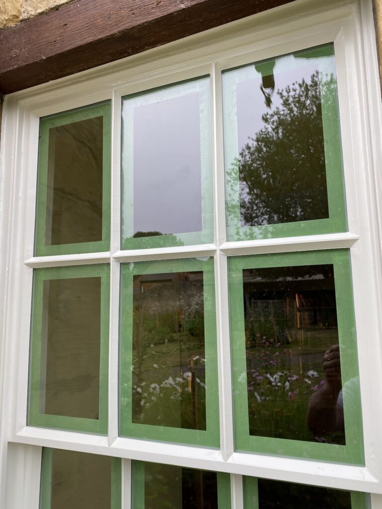 A window with white frames, typical of sash windows in Cambridge homes, has green painter’s tape outlining the glass panes; reflections of a garden and a person are visible.