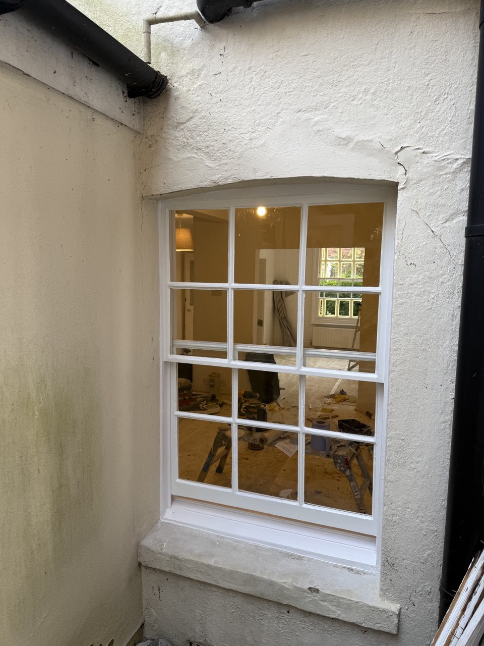 A white-framed sash window set in a light-coloured exterior wall, reflecting an indoor room under renovation with tools and materials scattered on the floor—typical of many sash windows Cambridge homes.