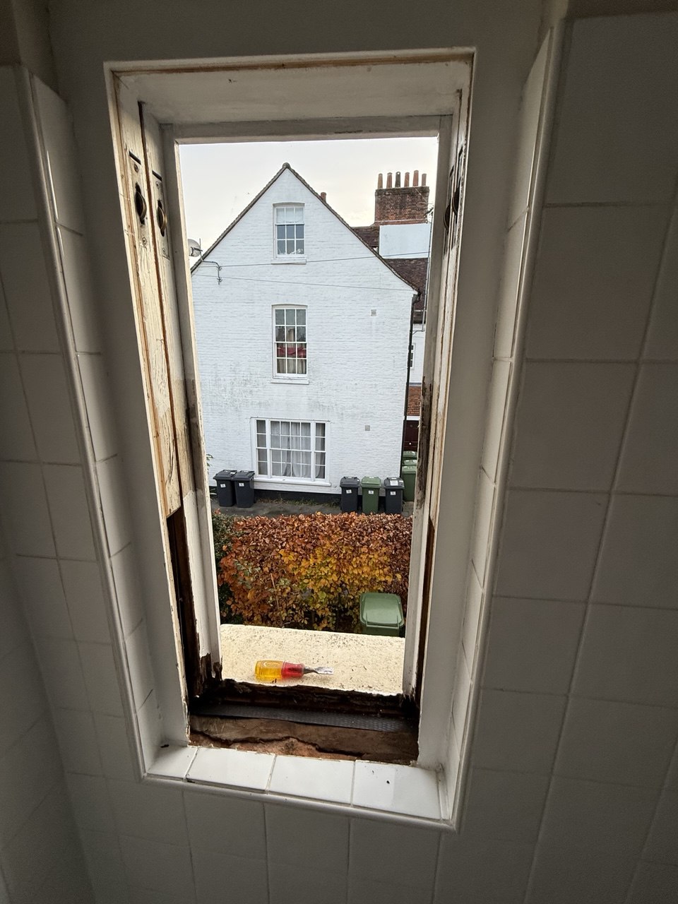 View through narrow sash windows in Cambridge with peeling paint, showing a white house, bins, and autumnal foliage outside. A plastic item sits on the windowsill.
